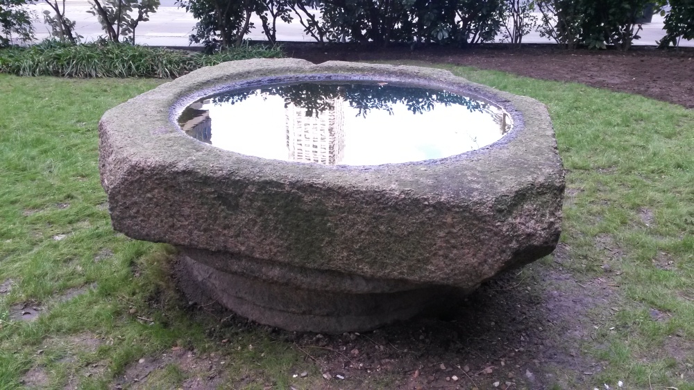 A large structure, possibily a font or column base, stands on a grassed area.  The grass is worn and muddy in places.  A round indent in the top of the structure is filled with water and a tower is reflected in the water.