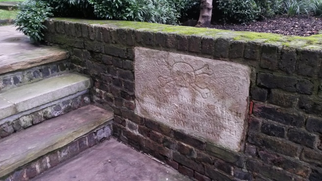 Image shows three steps on the left hand side, with a low wall on the right, within which is set an old, worn white stone plaque with unclear writing and a skull and crossbones carving